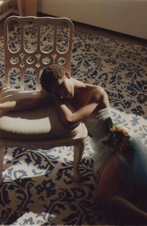 Woman in a light blue dress and floral accent, resting on a white decorative chair, on a blue and white patterned rug in sunlight.
