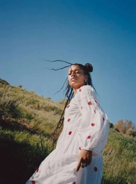 A woman in a white dress with red polka dots stands on a grassy hill under a clear blue sky, with her hair blowing in the wind.