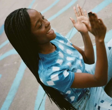 Smiling Black woman with braided hair is clapping in a blue tie-dye T-shirt and jeans against a blue and grey background outdoors.