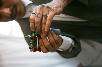Close-up of adult male tattoo artist holding tattoo gun, wearing grey suit jacket, in an indoor setting.
