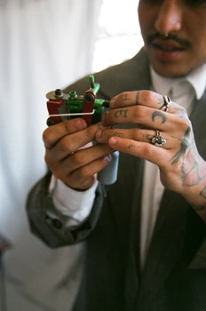 Close-up of adult male tattoo artist holding tattoo gun, wearing grey suit jacket, in an indoor setting.