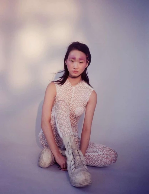 Asian woman with light make up, wearing lace patterned top, sitting down with white boots against gradient blue and white background.