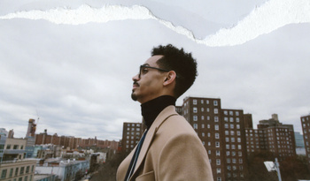 Man wearing a camel coat and black turtleneck standing against an urban rooftop and cloudy sky background