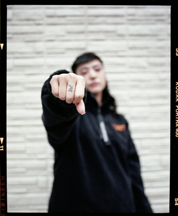 Woman with short dark hair gives a thumbs down gesture in front of a textured gray wall, wearing a black jacket and light pink nail polish.
