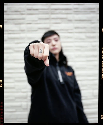 Woman with short dark hair gives a thumbs down gesture in front of a textured gray wall, wearing a black jacket and light pink nail polish.