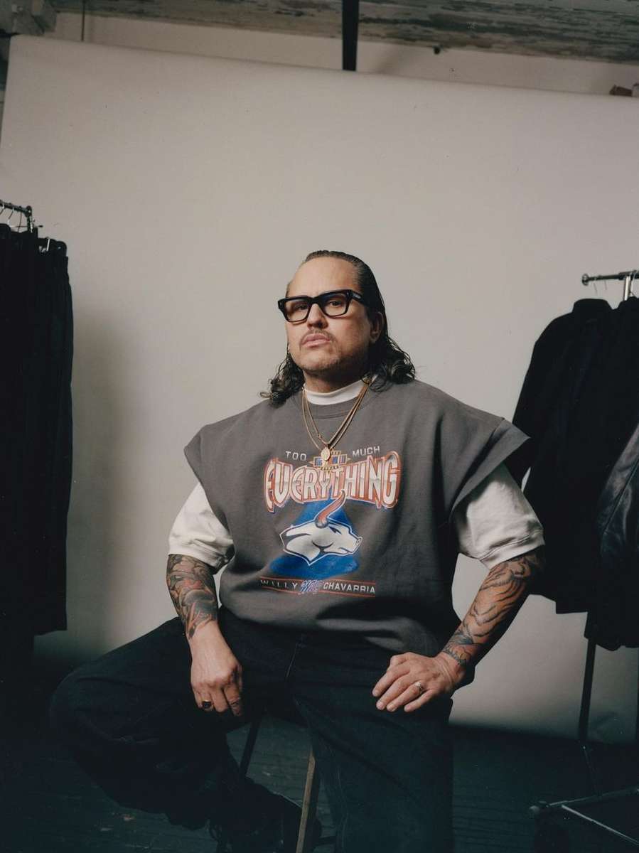 Portrait of adult man with long hair and glasses, wearing a graphic t-shirt while sitting on a stool in a photo studio.