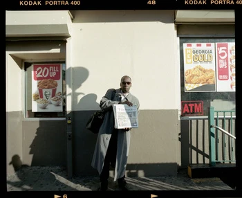An African-American man in a grey coat standing on a city street holding a newspaper with KFC and Georgia Gold ads behind him.