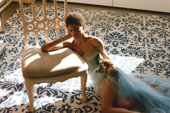Adult woman poses in a light blue tulle dress with floral accents, leaning on a white chair set on a blue patterned rug indoors.