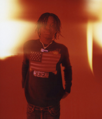 A young adult male with dreadlocks wearing a graphic sweater with an American flag stands in a warm-toned, indoor setting.