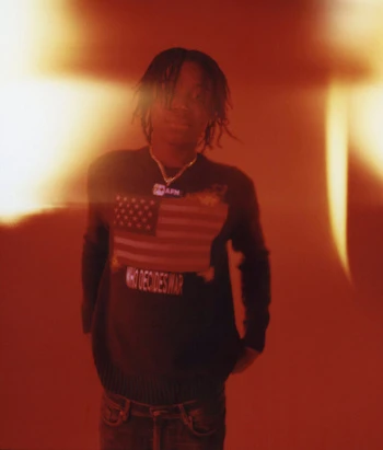 A young adult male with dreadlocks wearing a graphic sweater with an American flag stands in a warm-toned, indoor setting.