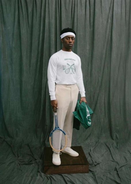 An African-American man stands on a wooden box in front of a green backdrop, holding a tennis racket and bag, wearing a white tennis outfit.