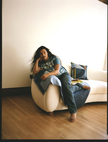 Adult female with curly hair sits on a couch with a patterned blanket indoors on a wood floor with a neutral wall in the background.