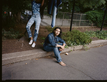 Two people modeling denim clothing outdoors; one man sitting barefoot on the edge of a curb, woman standing behind with jeans hanging nearby.