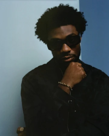 Portrait of a black man wearing dark sunglasses and a black shirt against a blue backdrop by photographer Levi Walton.