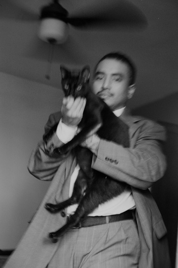 A man in a suit holds a black cat in an indoor setting with a ceiling fan in the background, captured in black and white photography.