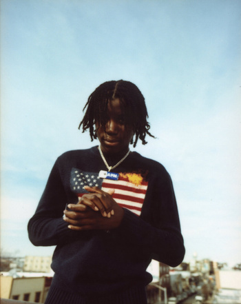 Portrait of a young adult male with dark hair wearing a Ralph Lauren sweater with an American flag design standing outdoors.