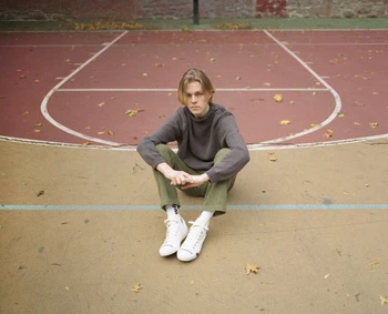 Young adult with blond hair sitting cross-legged on outdoor basketball court wearing a grey sweater, green pants, and white sneakers.