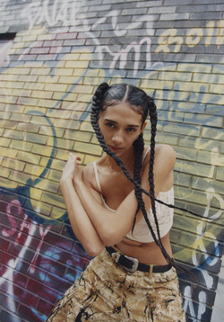 A woman with braided pigtails poses in front of graffiti covered brick wall wearing patterned pants and a cropped top in a medium shot.