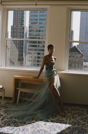 Model in a pale blue strapless tulle gown seated by a large window with city skyscrapers