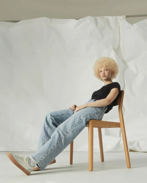 Woman with blonde afro sits on modern wood chair. She wears a black top, jeans, and sneakers, posing in a minimalist studio with white background.