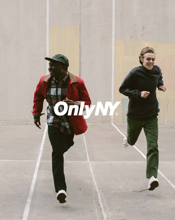 Two young adults are running outdoors on a light-grey concrete surface. One wears a red jacket, the other a black sweater. Large logo visible.