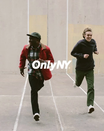 Two young adults are running outdoors on a light-grey concrete surface. One wears a red jacket, the other a black sweater. Large logo visible.