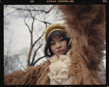 Portrait of an Asian woman wearing a yellow beret and ruffled shirt, posing with a brown faux fur coat in an outdoor environment with bare trees.