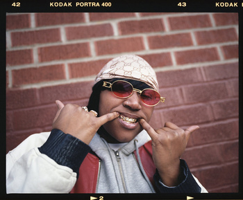 Portrait of an adult female with gold teeth wearing red glasses, a durag, and a grey hooded sweatshirt against a red brick wall.