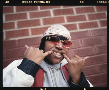Portrait of an adult female with gold teeth wearing red glasses, a durag, and a grey hooded sweatshirt against a red brick wall.