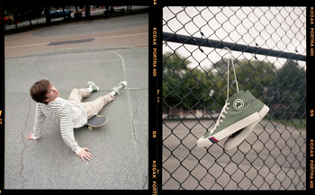 A young man sits on a skateboard in a casual urban setting. Sneakers hang on a chain-link fence with green trees in the background.
