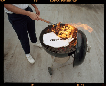 Overhead shot of a man grilling with flames rising from a grill, featuring hotdogs and a VICELAND-branded shirt.