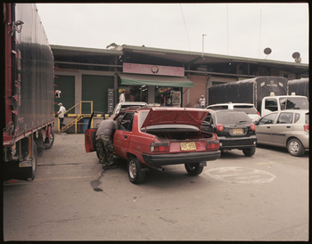 Red car with open trunk at warehouse loading dock with a person attending to it, surrounded by trucks and other vehicles.