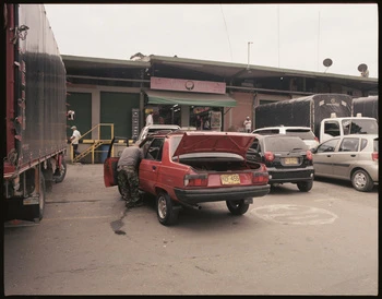 Red car with open trunk at warehouse loading dock with a person attending to it, surrounded by trucks and other vehicles.