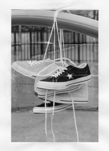 Two pairs of converse sneakers are hung by their laces from a metal bar in a grayscale photograph with an out-of-focus background.