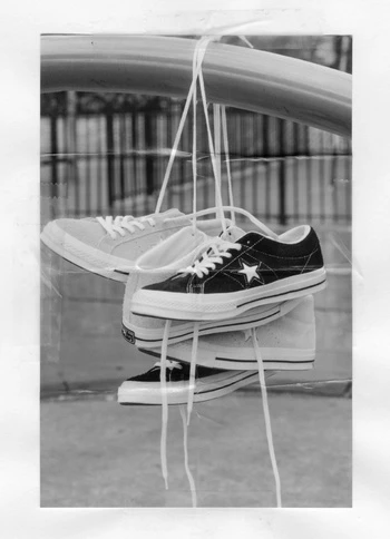 Two pairs of converse sneakers are hung by their laces from a metal bar in a grayscale photograph with an out-of-focus background.