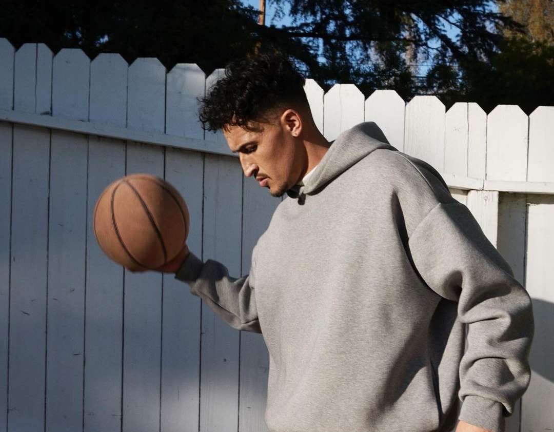 Young adult male in gray hoodie holding a basketball outdoors against a white wooden fence on a sunny day with visible tree branches.