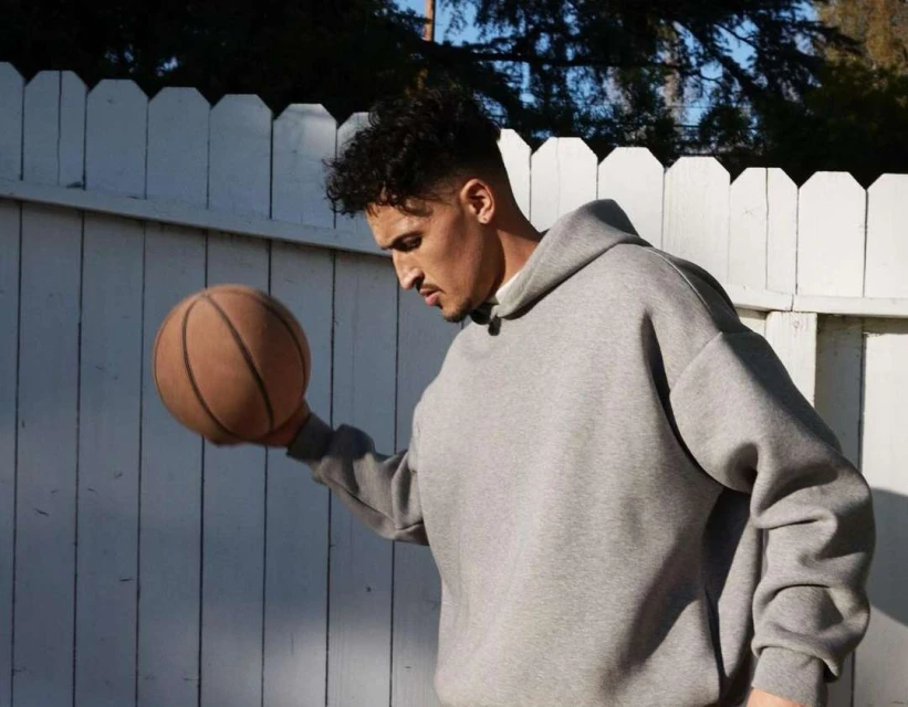 Young adult male in gray hoodie holding a basketball outdoors against a white wooden fence on a sunny day with visible tree branches.
