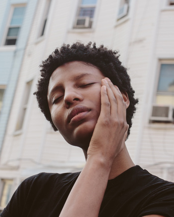 Close-up portrait of a Black adult woman with closed eyes and hand near face, posing outdoors with an urban building background.