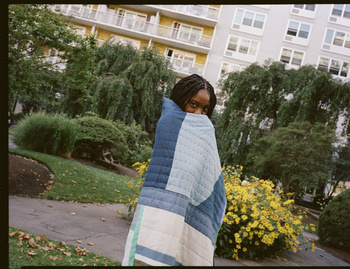 Portrait of an African-American woman wrapped in a blue patchwork quilt in an urban green space with yellow flowers.