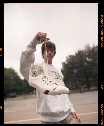 Low-angle shot of person holding white high-top sneaker with red and blue stripes against an outdoor court and tree-lined background.
