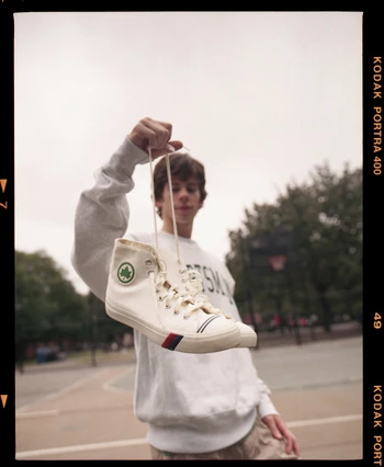 Low-angle shot of person holding white high-top sneaker with red and blue stripes against an outdoor court and tree-lined background.