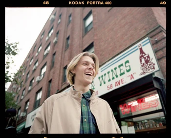 Smiling young man in a jacket stands outside a wineshop with a brick building backdrop on a cloudy day, creating an urban scene.