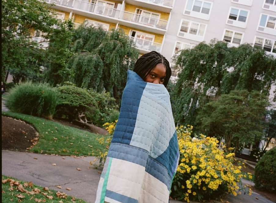 Young adult woman with dark hair wrapped in a blue patchwork quilt outdoors in a garden setting with apartment buildings.