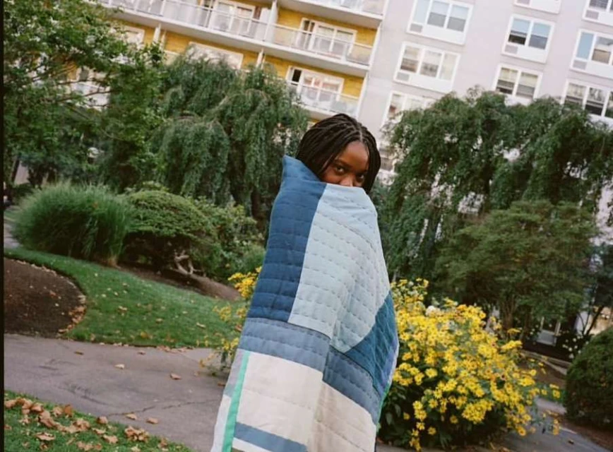 Young adult woman with dark hair wrapped in a blue patchwork quilt outdoors in a garden setting with apartment buildings.