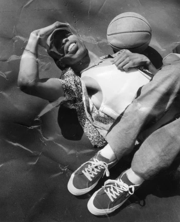 Black and white portrait of a young adult male with a basketball, wearing a cap, tank top, and sneakers, posing on a crinkled surface.