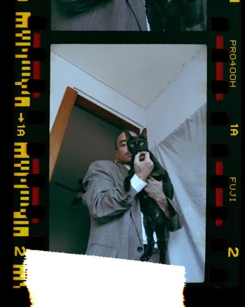 Adult male in grey suit holding a black cat with film border. The cat's eyes are green; the man has an earring and tattoos.
