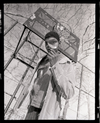 Portrait of an African-American adult male holding a magnifying glass to his eye in front of a basketball court and chainlink fence.