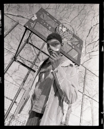 Portrait of an African-American adult male holding a magnifying glass to his eye in front of a basketball court and chainlink fence.