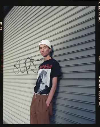 A young man wearing a white beanie and a black T-shirt with a graphic print stands in front of a gray metal rolling shutter door with graffiti.