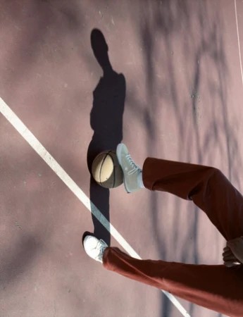 Legs of a person wearing rust red trousers and white sneakers resting foot on a basketball on a paved court with long shadow